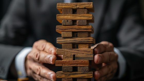 A man in a suit meticulously moves a wooden block in a Jenga tower, strategically placed on a table surrounded by coins